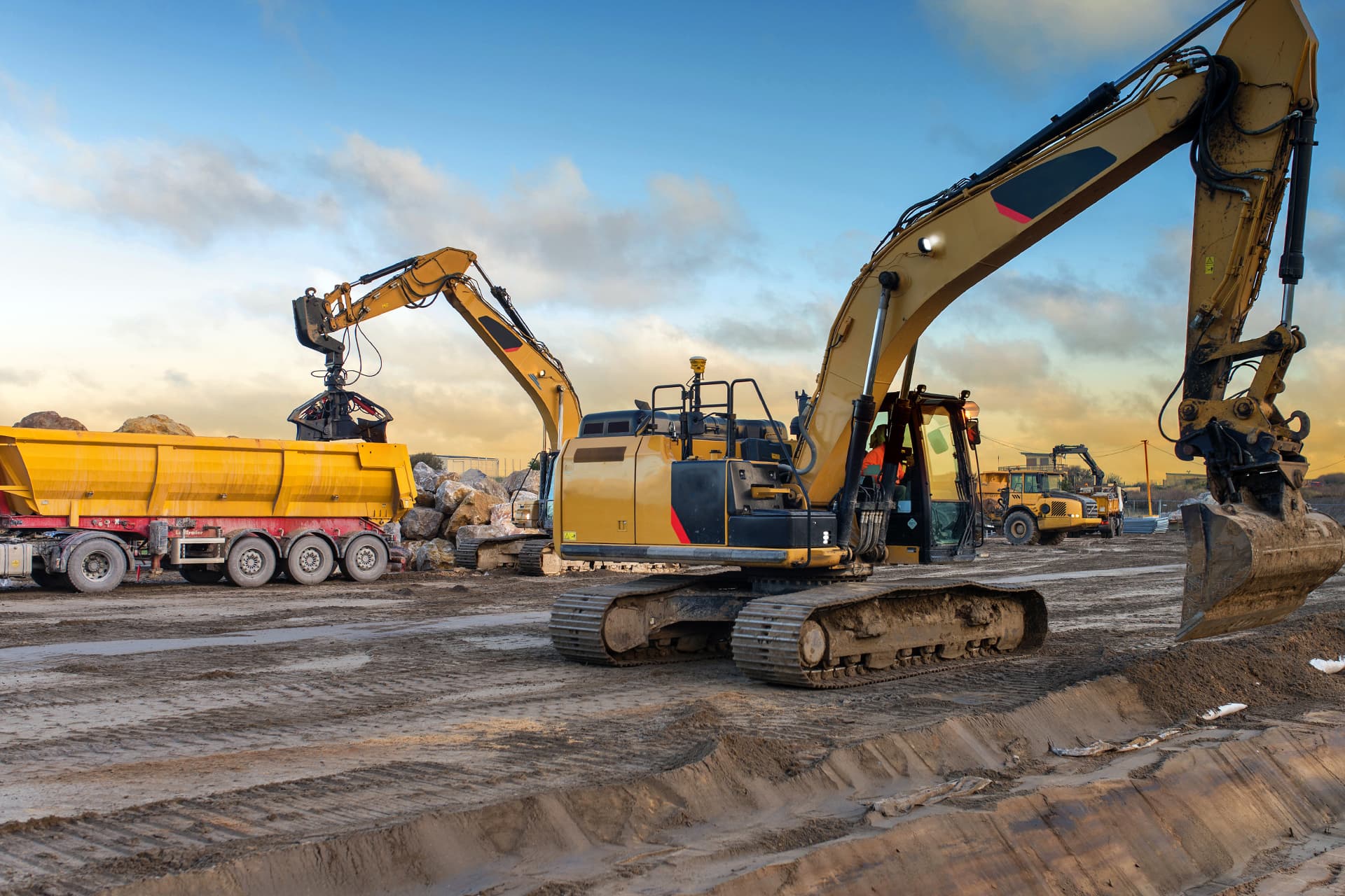 Large yellow excavators operate at an outdoor construction site, one loading rocks into a yellow dump truck while other heavy machinery works in the background on muddy, graded ground under a partly cloudy sky.